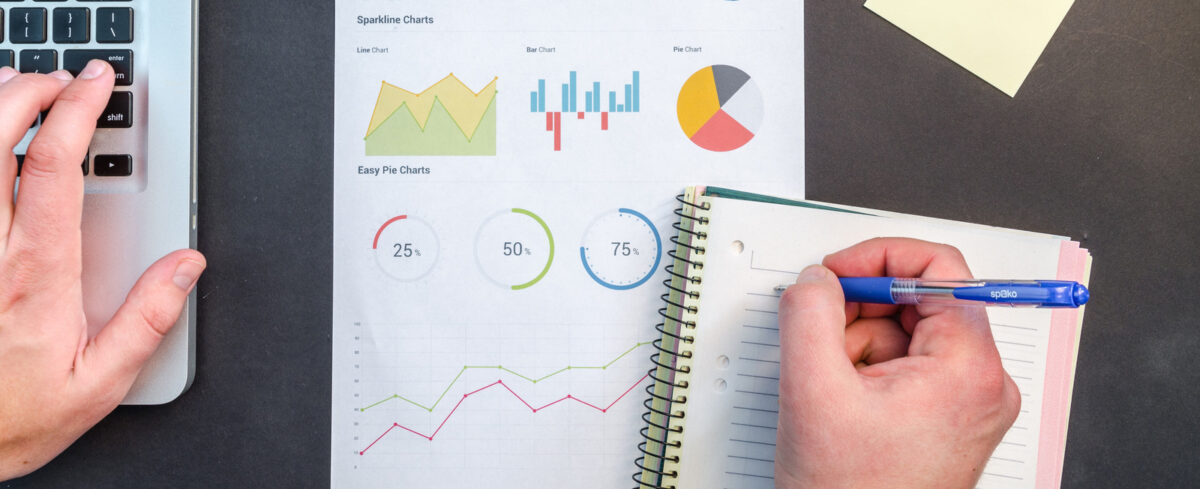 person writing in notebook with computer and graphs